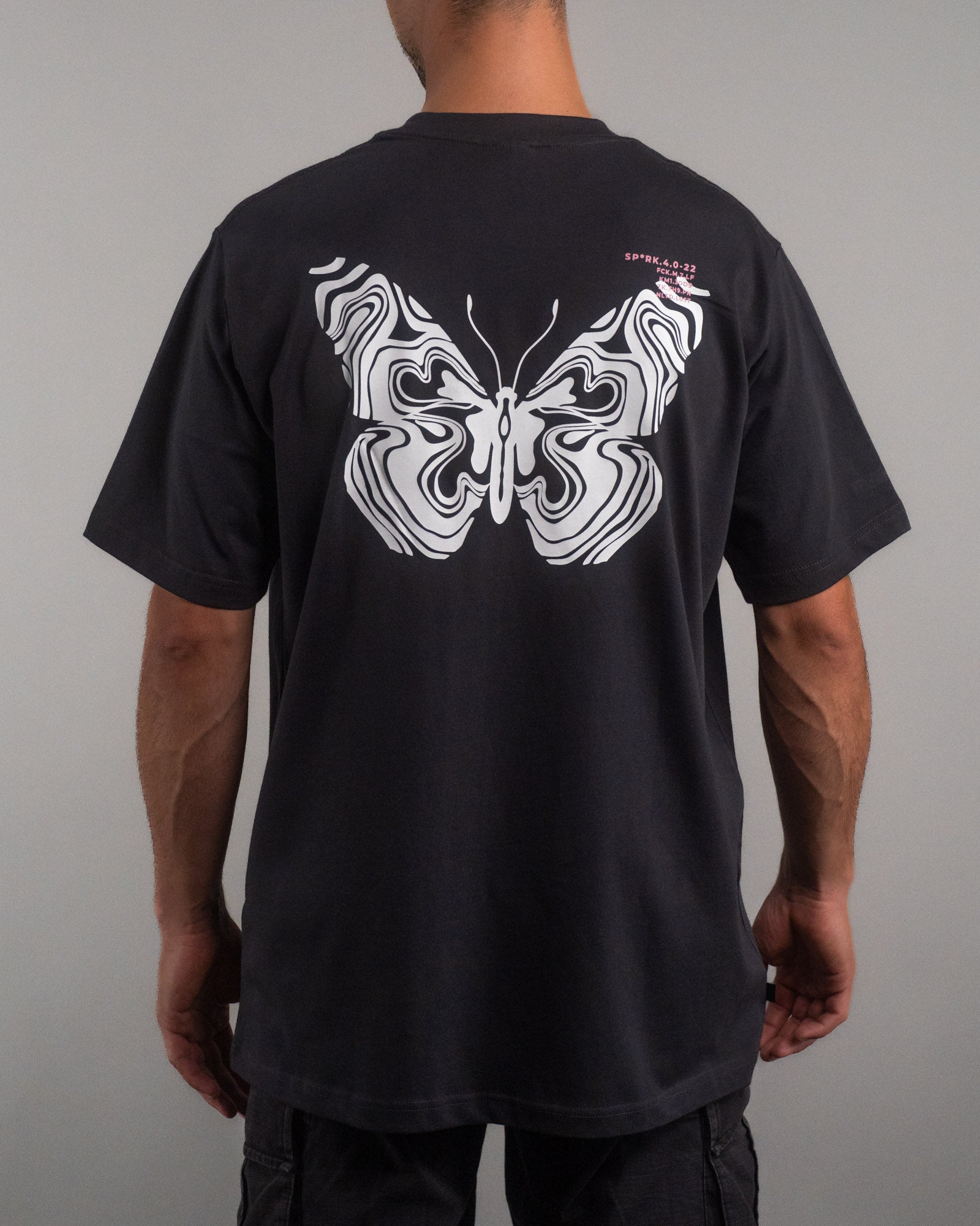 Rückenansicht desselben schwarzen T-Shirts mit großflächigem, weißem Schmetterlingsprint in marmorierter Liniengrafik. Rechts oben ist ein kleiner rosa Textprint mit Zahlen- und Buchstabenkombinationen sichtbar.