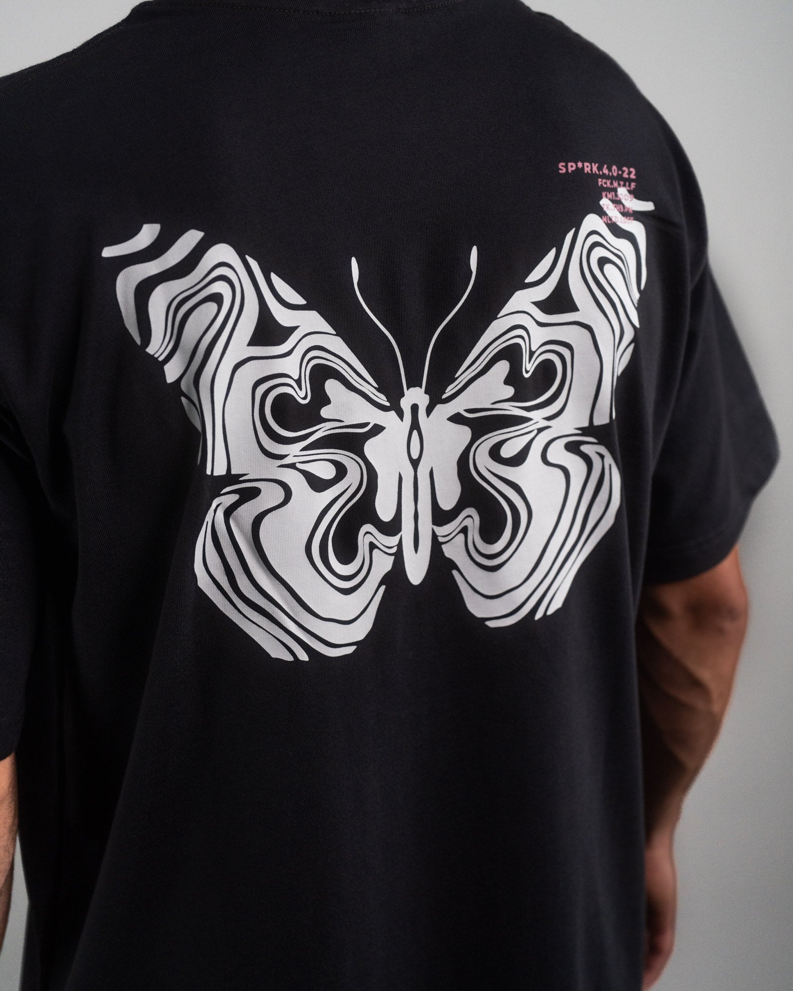 Detailansicht eines pinken Labelpatches am unteren Saum des T-Shirts. Der Patch ist mit schwarzer Schrift bedruckt und wird von einer auffälligen blauen Ziernaht durchzogen.