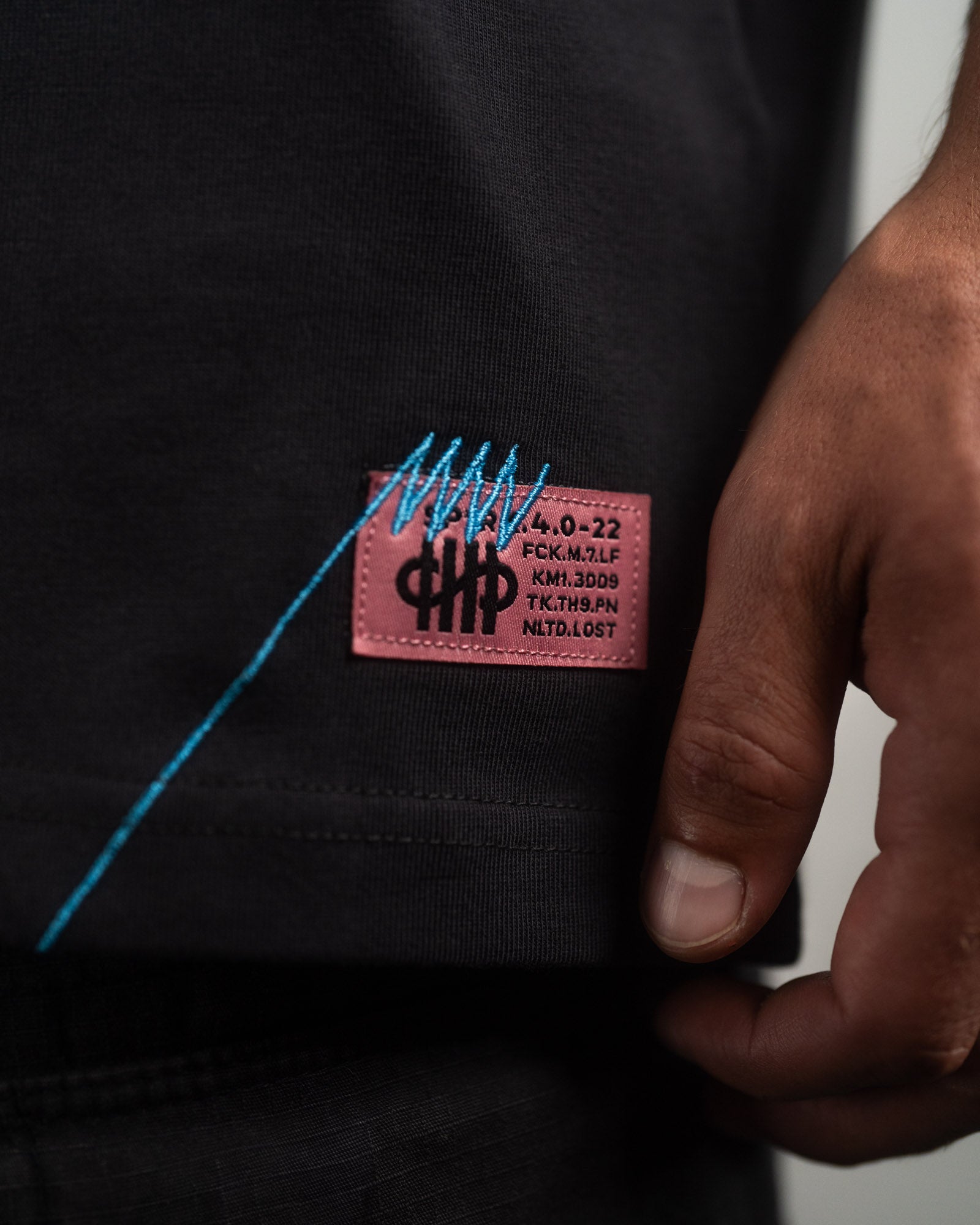 Nahaufnahme des seitlichen Labels am Saum des T-Shirts. Auf dem schwarzen Etikett steht in Weiß „SP*RK“.