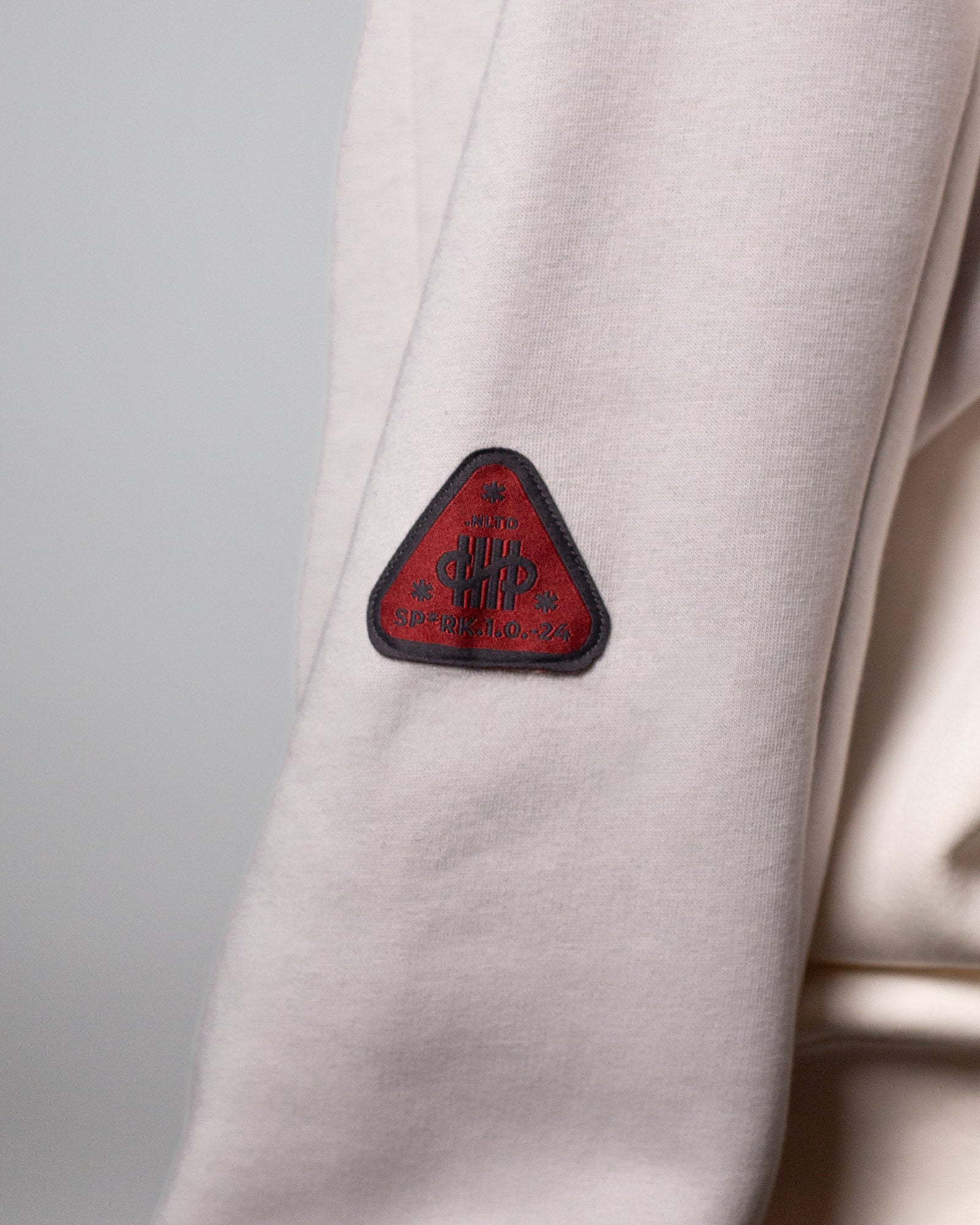 Nahaufnahme des linken Ärmels eines cremefarbenen Hoodies mit grauen Ärmeln. Auf dem Oberarm ist ein rotes, dreieckiges Patch mit schwarzer Umrandung aufgenäht, das grafische und typografische Elemente zeigt.