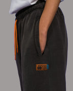 Seitliche Nahaufnahme der schwarzen Jogginghose mit sichtbarer Hosentasche. Darunter ist ein rechteckiges Lederpatch mit aufgesticktem Logo, Zahlen und Text in Schwarz und Blau angebracht. Die orangefarbene Kordel ist teilweise sichtbar.