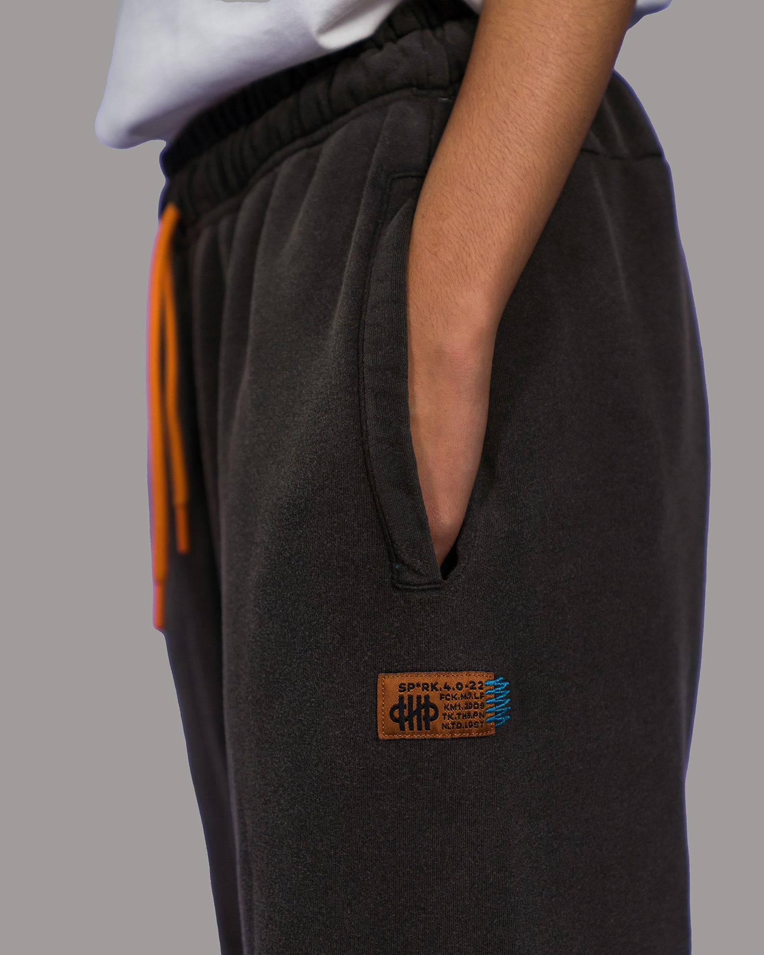 Seitliche Nahaufnahme der schwarzen Jogginghose mit sichtbarer Hosentasche. Darunter ist ein rechteckiges Lederpatch mit aufgesticktem Logo, Zahlen und Text in Schwarz und Blau angebracht. Die orangefarbene Kordel ist teilweise sichtbar.