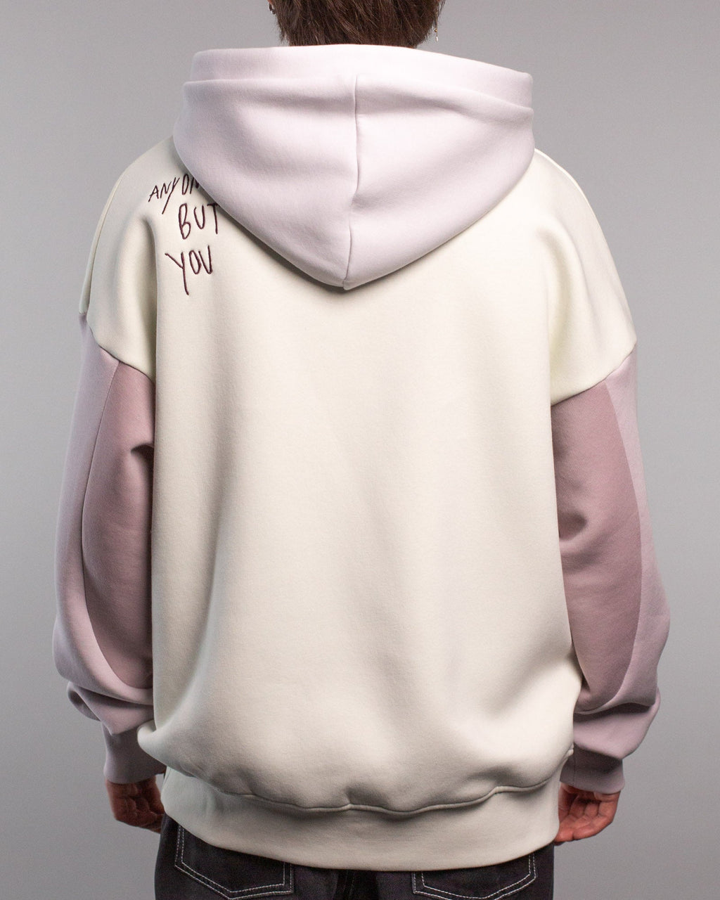 Rückansicht des Hoodies mit Schriftzug „ANYONE BUT YOU“ in dunkelvioletter Stickerei auf der linken Schulter. Die Kapuze ist fliederfarben, Rumpf und Rücken des Hoodies sind cremefarben gehalten.