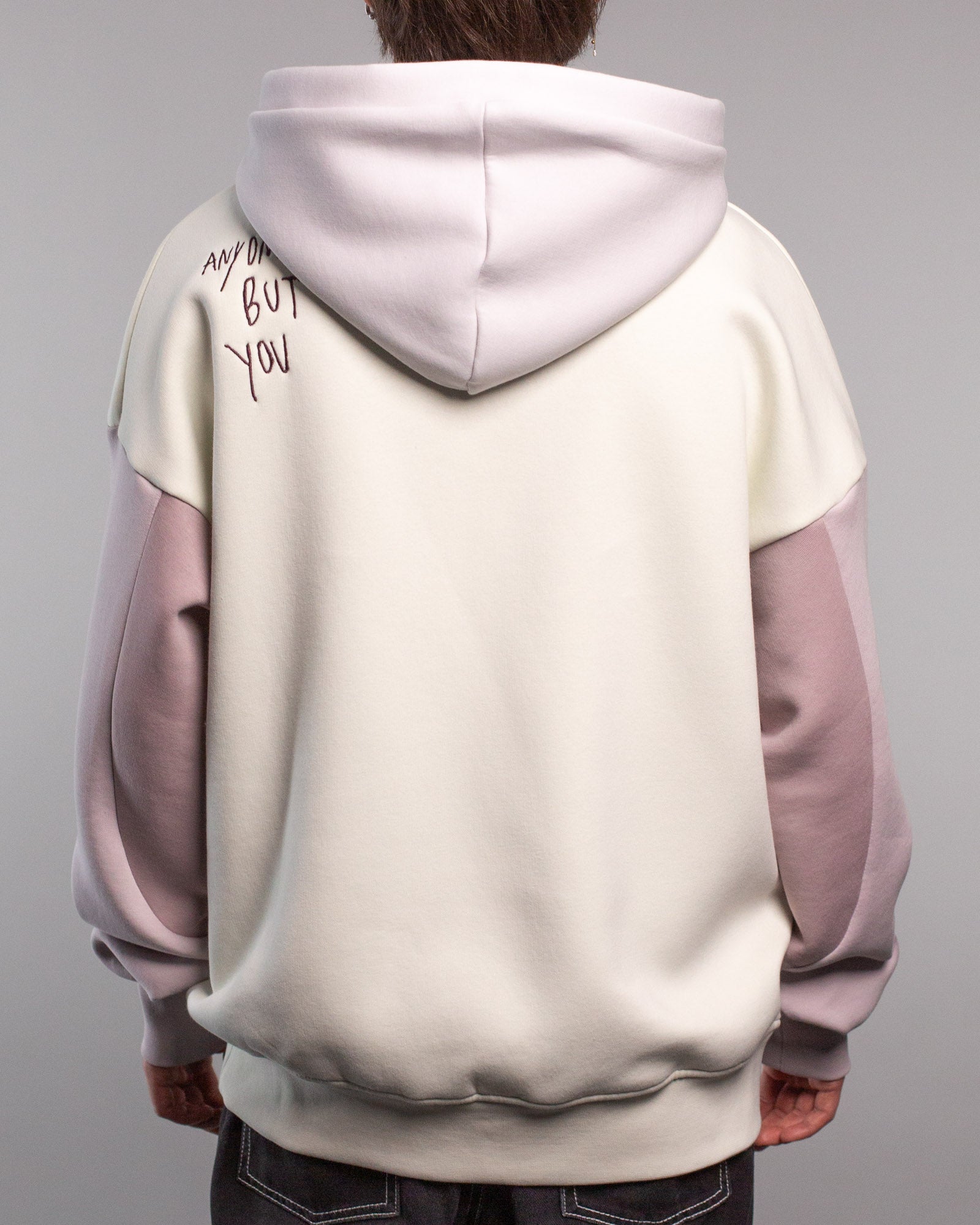 Rückansicht des Hoodies mit Schriftzug „ANYONE BUT YOU“ in dunkelvioletter Stickerei auf der linken Schulter. Die Kapuze ist fliederfarben, Rumpf und Rücken des Hoodies sind cremefarben gehalten.