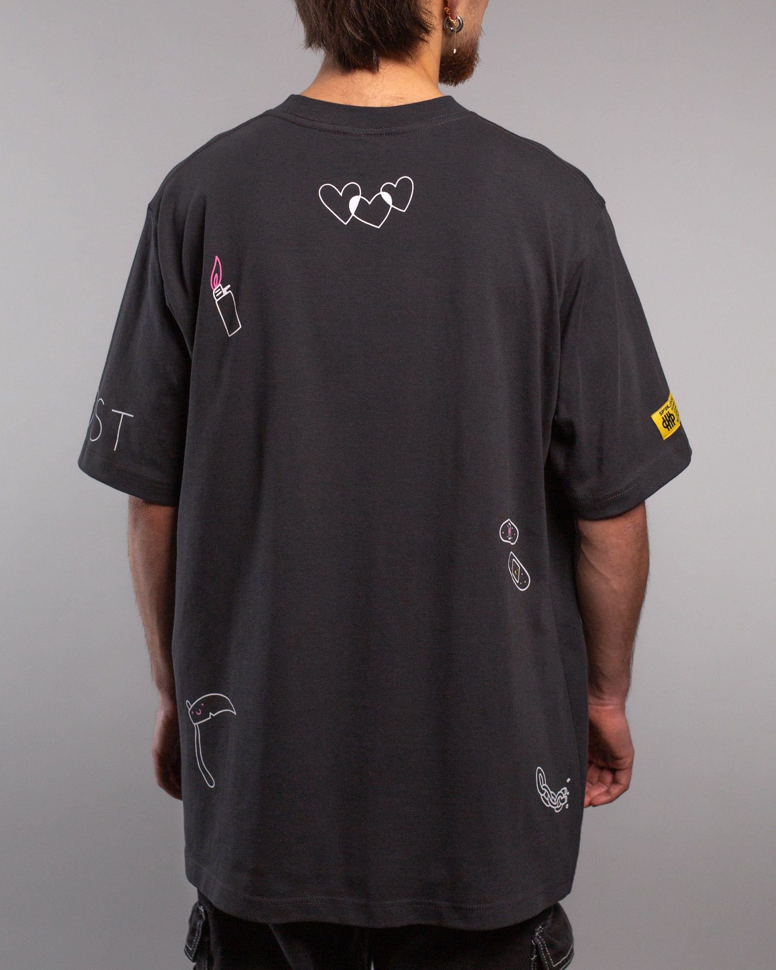 Rückansicht des T-Shirts mit mehreren kleinen Illustrationen: drei sich überschneidende weiße Herzen im oberen Rückenbereich, ein pinkes Feuerzeug links, ein lachender Sensenmann rechts unten und weitere kleine Symbole wie Pflaster, Ketten und ein gelbes Label an der rechten Ärmelseite.