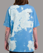 Rückansicht eines blau-weißen Batikshirts mit wolkenartigem Muster. Auf dem oberen Rücken befindet sich ein schwarzer Textprint in versetzter Zeilenstruktur. Das Muster zieht sich unregelmäßig über die gesamte Fläche.