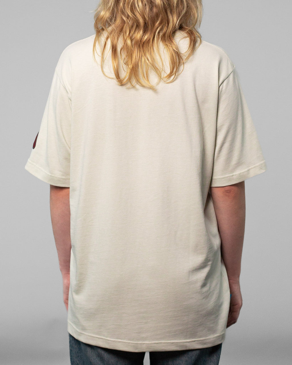 Rückansicht eines beigefarbenen T-Shirts ohne Aufdruck. Das Shirt hat eine lockere Passform mit geradem Saum, kurzen Ärmeln und Rundhalsausschnitt. Der Fokus liegt auf der schlichten Rückseite und der weichen Stoffstruktur. Der Hintergrund ist neutral grau gehalten.