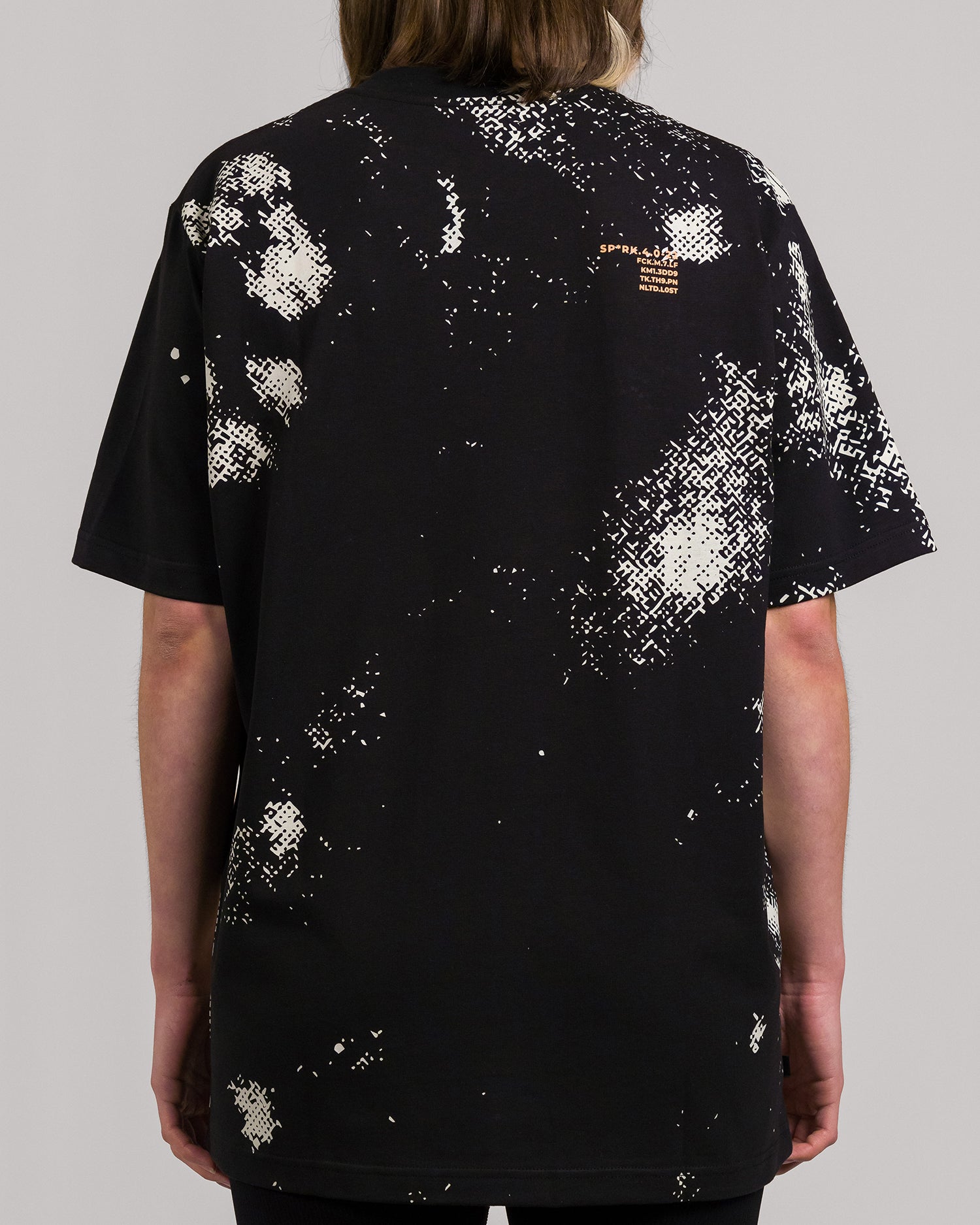Rückansicht desselben schwarzen Oversize-T-Shirts mit verstreutem beigen und weißem Pixelmuster. Im oberen rechten Bereich befindet sich ein kleiner gelboranger Textdruck mit Zahlen- und Buchstabenkombinationen. Die übrige Fläche ist überwiegend schwarz mit dezentem Musterverlauf.