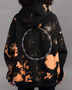 Die Rückseite des Hoodies zeigt ein durchgehendes Rastermuster in Orange auf schwarzem Stoff. Im oberen Bereich rechts ist ein weißer Schriftzug mit alphanumerischem Code aufgedruckt. Zentral im Rückenbereich befindet sich ein großer, runder Druck aus weißen Stacheldraht-Elementen, die zu einem Kreis verbunden sind.