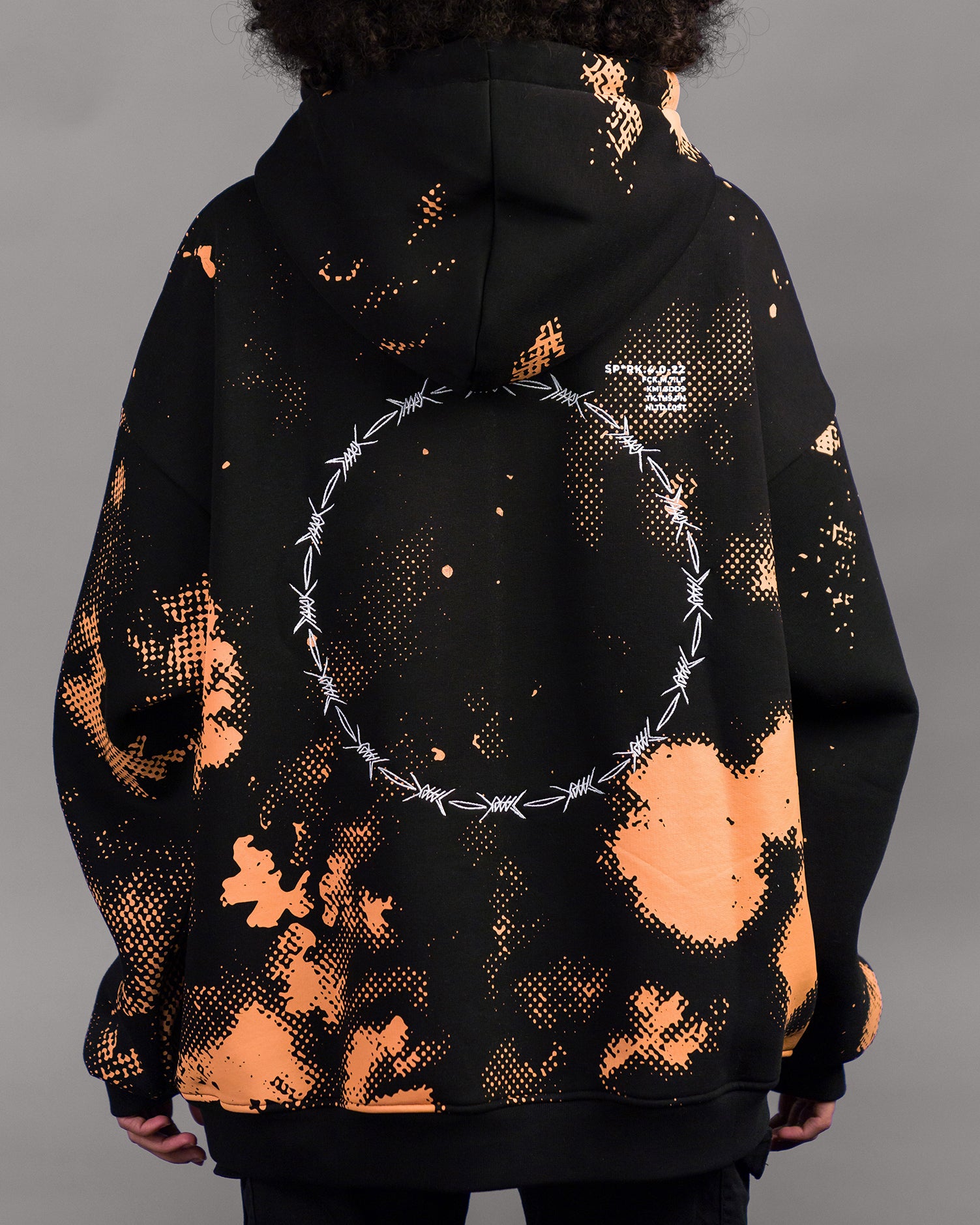 Die Rückseite des Hoodies zeigt ein durchgehendes Rastermuster in Orange auf schwarzem Stoff. Im oberen Bereich rechts ist ein weißer Schriftzug mit alphanumerischem Code aufgedruckt. Zentral im Rückenbereich befindet sich ein großer, runder Druck aus weißen Stacheldraht-Elementen, die zu einem Kreis verbunden sind.