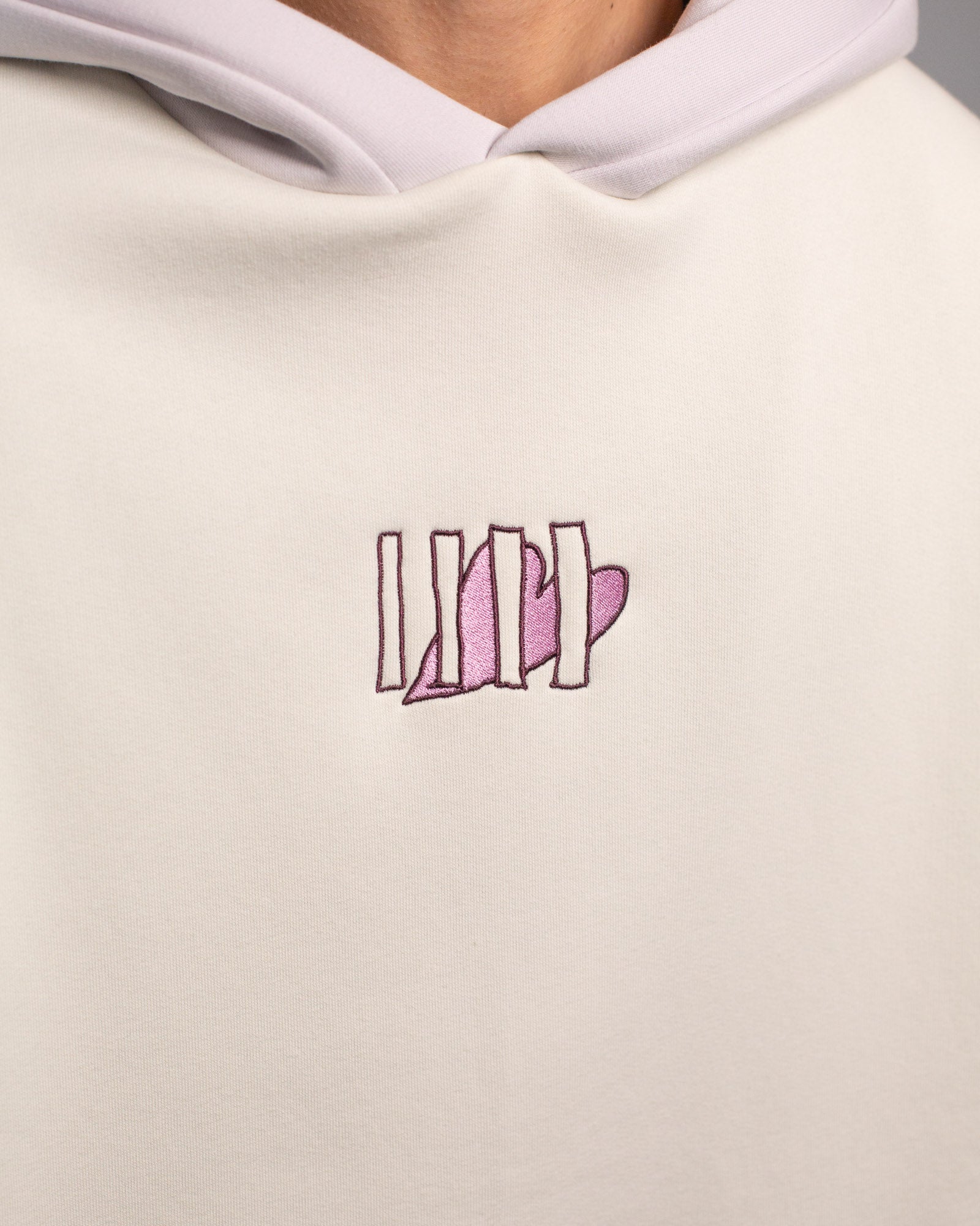  Detailansicht des Brustmotivs auf dem Hoodie. Zu sehen ist ein stilisiertes Herz in glänzendem Rosa, das von fünf vertikalen Linien in dunkler Stickerei überlagert wird.