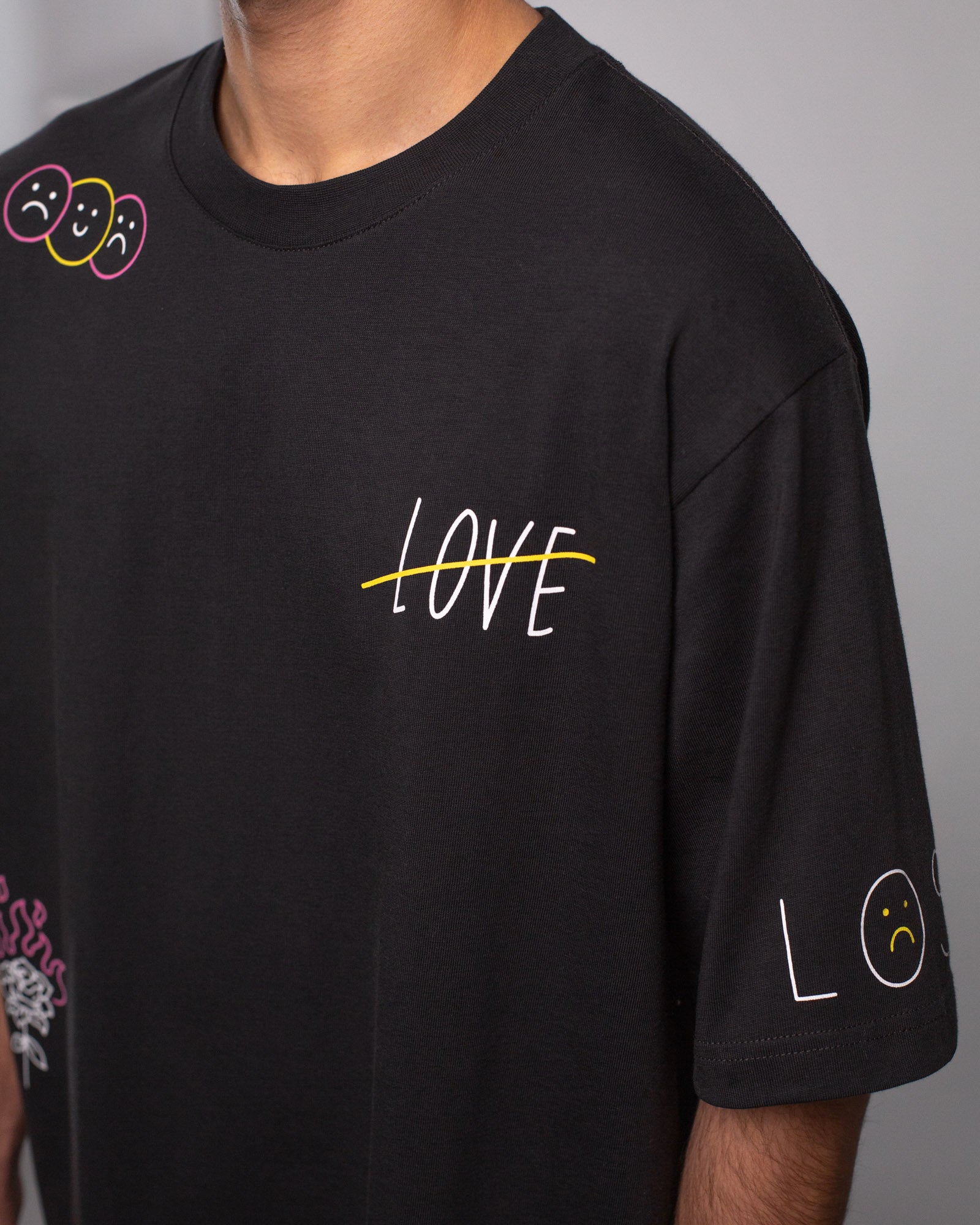 Seitliche Nahaufnahme des T-Shirts mit Fokus auf das durchgestrichene Wort „LOVE“ in Weiß mit gelber Linie, sowie dem Schriftzug „LOST“ auf dem rechten Ärmel mit traurigem gelbem Smiley als Buchstabe „O“.