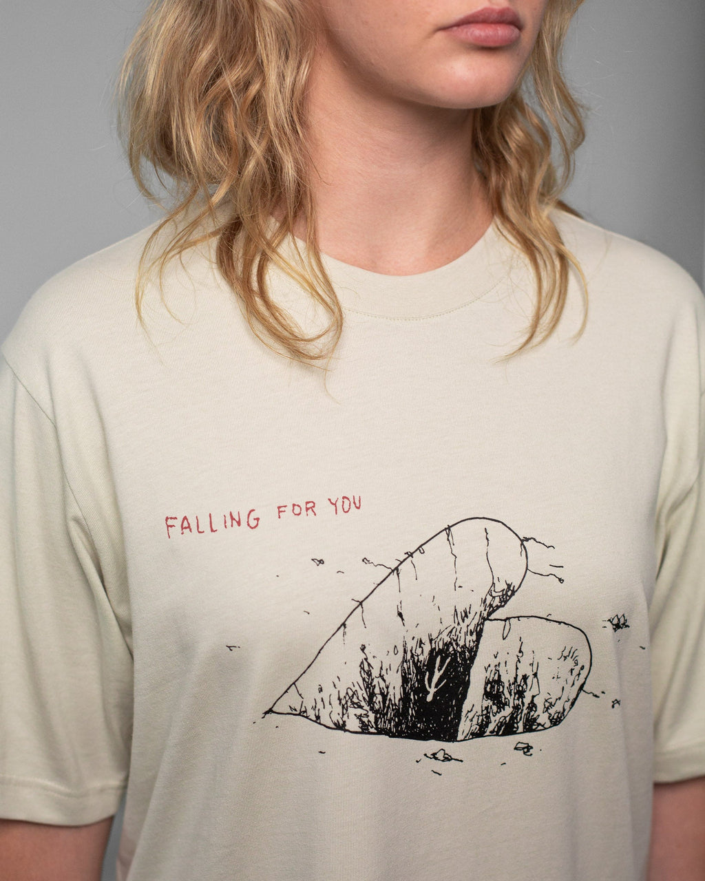 Detailaufnahme der Vorderseite eines beigefarbenen T-Shirts mit grafischem Print. Zu sehen ist ein schwarzes, halbiertes Herz mit feinen Linien und Strukturen, die an Risse oder Wurzeln erinnern. Darüber steht der rote Schriftzug „FALLING FOR YOU“. Der Fokus liegt auf dem illustrativen Motiv und der Textgestaltung auf der Brust.
