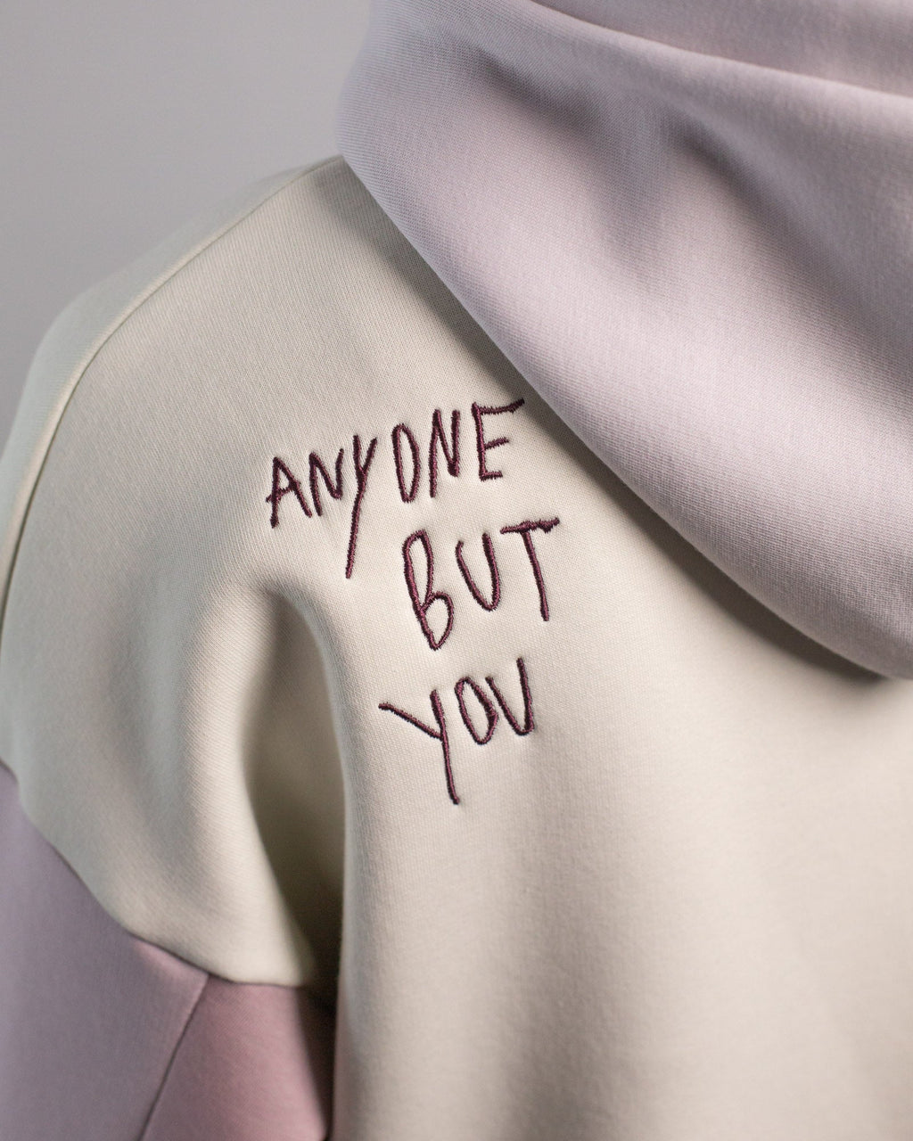 Detailaufnahme der linken Schulter des Hoodies von hinten. Die dunkelviolette Stickerei zeigt den handschriftlich anmutenden Schriftzug „ANYONE BUT YOU“ auf cremefarbenem Grund.