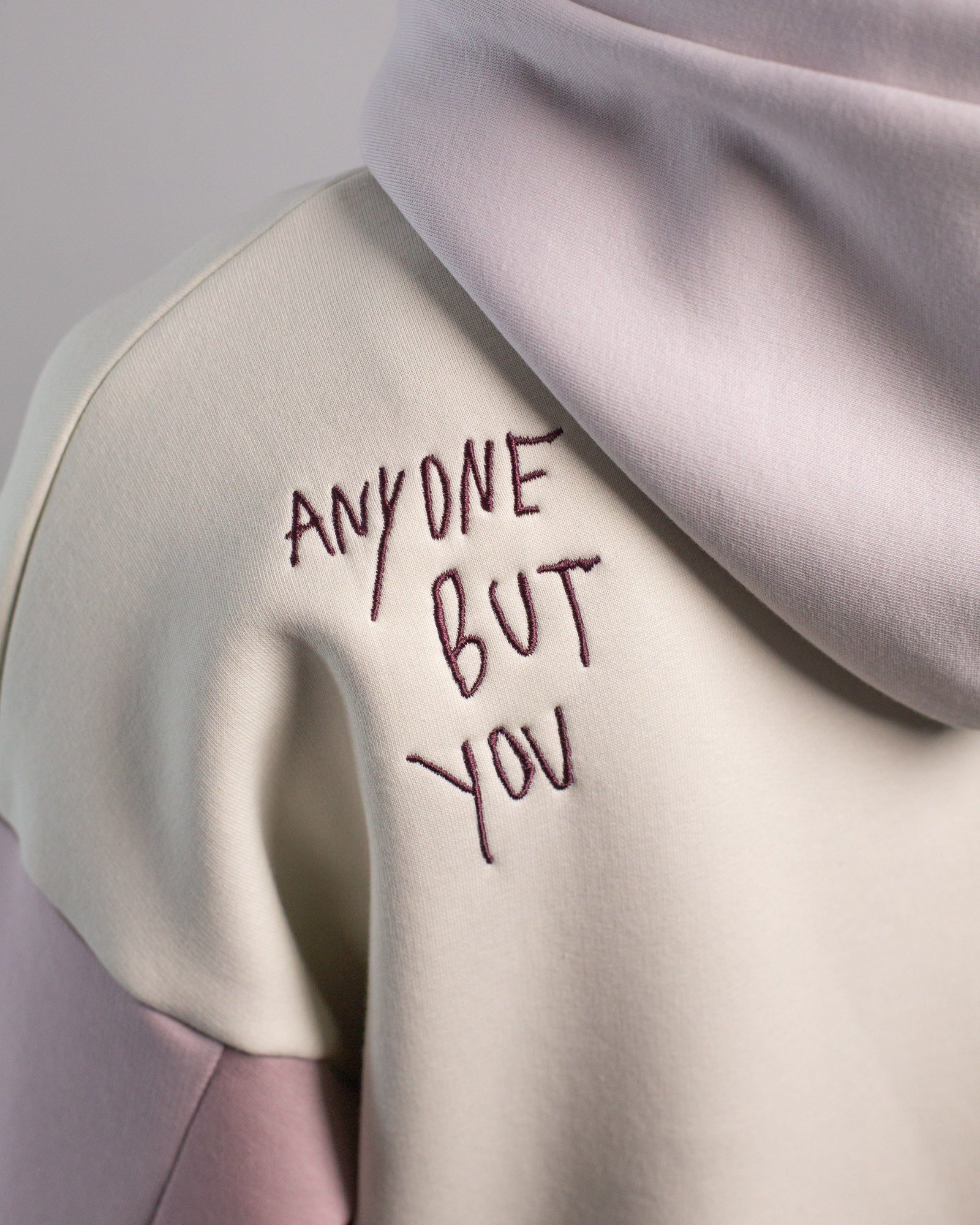 Detailaufnahme der linken Schulter des Hoodies von hinten. Die dunkelviolette Stickerei zeigt den handschriftlich anmutenden Schriftzug „ANYONE BUT YOU“ auf cremefarbenem Grund.