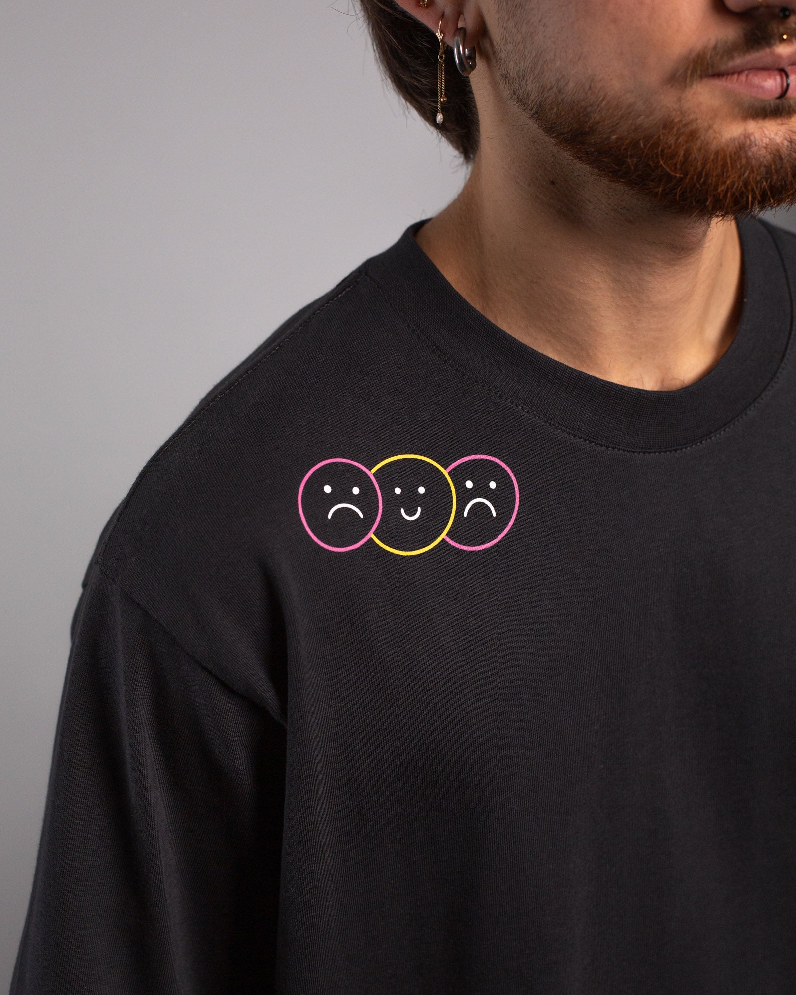 Detailansicht der linken Schulterpartie mit drei stilisierten Smiley-Gesichtern in rosa, gelb und rosa Kreisrahmen – zwei traurig, eines fröhlich in der Mitte.