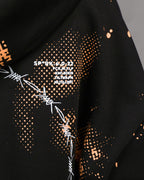 Nahaufnahme des weißen Stacheldraht-Kreisdrucks auf schwarzem Stoff mit orangefarbenem Punktraster. Rechts daneben befindet sich der weiße Textaufdruck „SP*RK 4.0.22“ sowie darunter mehrere Codezeilen in Großbuchstaben.