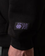 Nahaufnahme des Ärmelpatches unten rechts am Hoodie. Auf dem schwarzen Label befindet sich ein lilafarbenes Emblem mit stilisierter Zählgrafik sowie technische Anmutung durch kryptische Zeichen wie „SP*RK.I.O.-24“ und weitere Codezeilen.