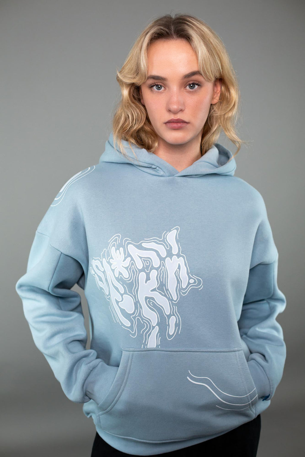 Eine Person trägt einen hellblauen Kapuzenpullover mit weißem, abstraktem Linienmotiv auf der Vorderseite. Der Print zeigt fließende, organische Formen auf Brusthöhe, ergänzt durch geschwungene Linien an der Kängurutasche. Der Hoodie hat eine weite Passform mit breiten Bündchen und einer weichen Stoffstruktur. Der Fokus liegt auf dem grafischen Frontdesign des Hoodies vor neutralem grauem Hintergrund.