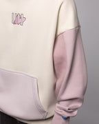 Detailansicht des linken Vorderteils des Hoodies mit Fokus auf das Zusammenspiel der Farbbereiche: cremefarbener Rumpf, fliederfarbene Tasche und Ärmel. Das Herz-Logo auf der Brust ist ebenfalls sichtbar.