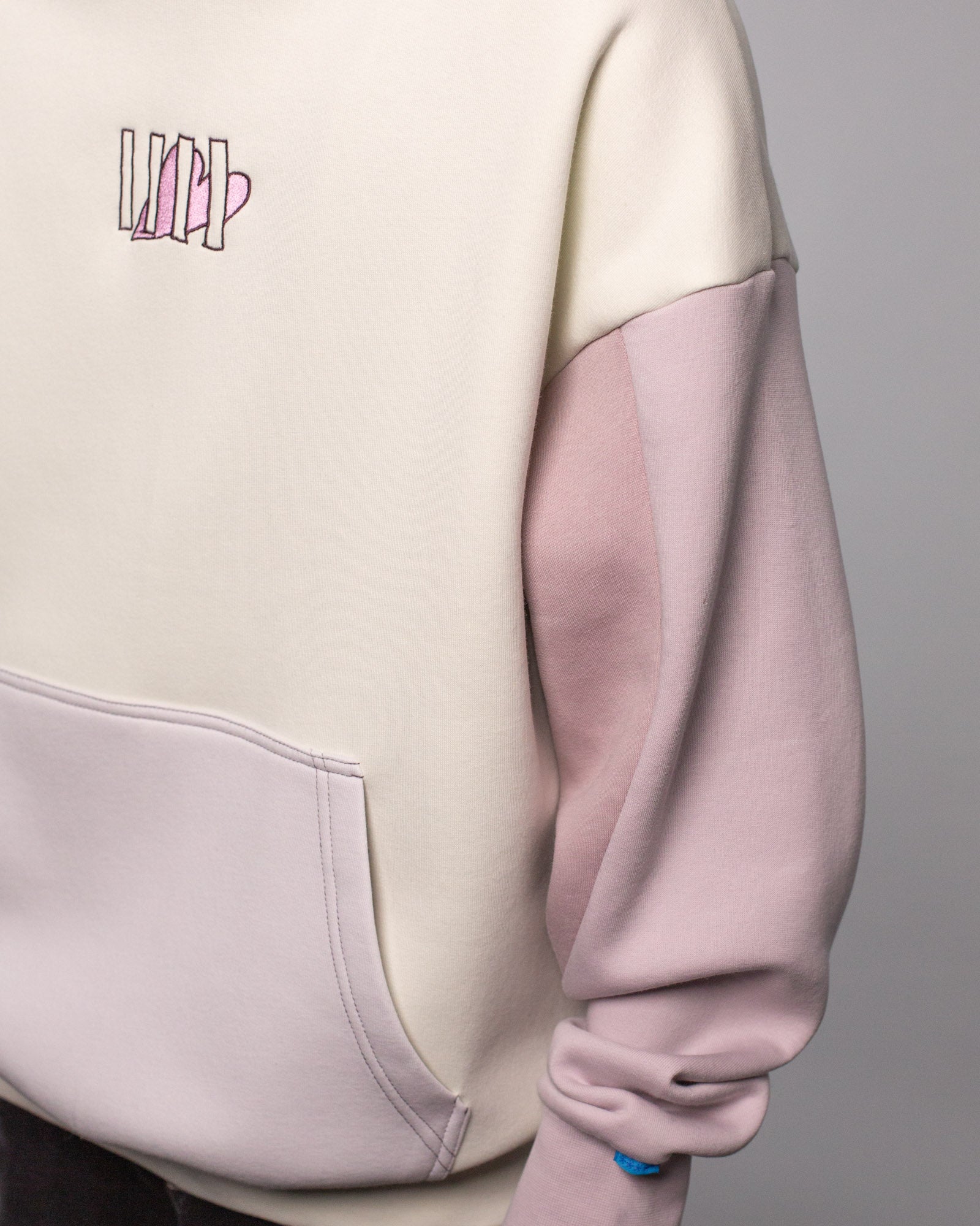 Detailansicht des linken Vorderteils des Hoodies mit Fokus auf das Zusammenspiel der Farbbereiche: cremefarbener Rumpf, fliederfarbene Tasche und Ärmel. Das Herz-Logo auf der Brust ist ebenfalls sichtbar.