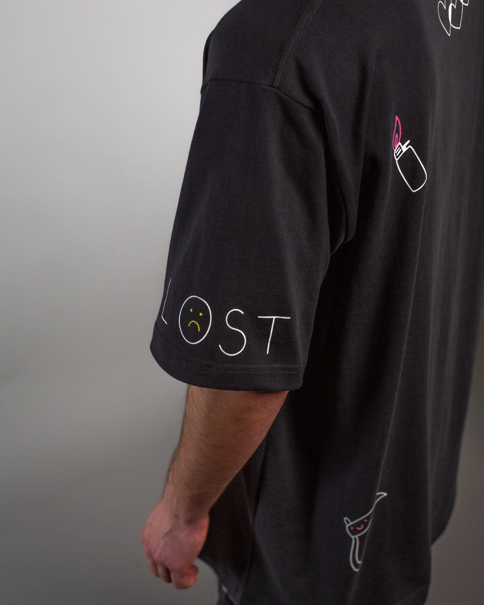 Seitliche Rückansicht mit Fokus auf die Illustration des Schriftzugs „LOST“ auf dem Ärmel sowie die darunter platzierte Sense mit freundlichem Gesicht, alles in weißen Linien auf schwarzem Stoff.