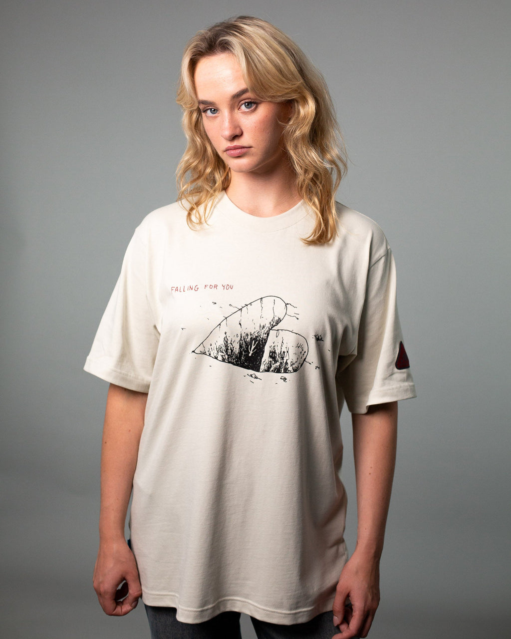 Eine Person trägt ein beigefarbenes T-Shirt mit schwarzem Grafikprint auf der Vorderseite. Das Motiv zeigt ein halbiertes Herz mit feinen Linien und Struktur, darüber steht der rote Schriftzug „FALLING FOR YOU“. Auf dem linken Ärmel befindet sich ein kleiner roter Patch in Dreiecksform. Das Shirt hat eine lockere Passform mit Rundhalsausschnitt. Der Fokus liegt auf dem illustrativen Frontdesign und den dezenten Designdetails.