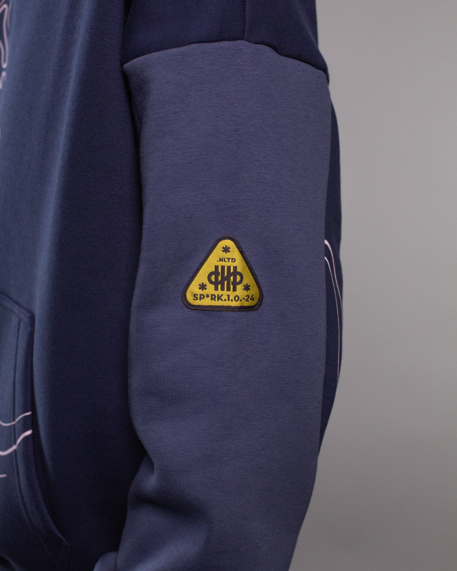Detailansicht des linken Ärmels mit gelb-schwarzem Labelpatch in Dreiecksform. Die Aufschrift „SP*RK 1.0.24“ sowie stilisierte Symbole erscheinen innerhalb des Patches auf dem abgesetzten Ärmel in dunklem Violettblau.