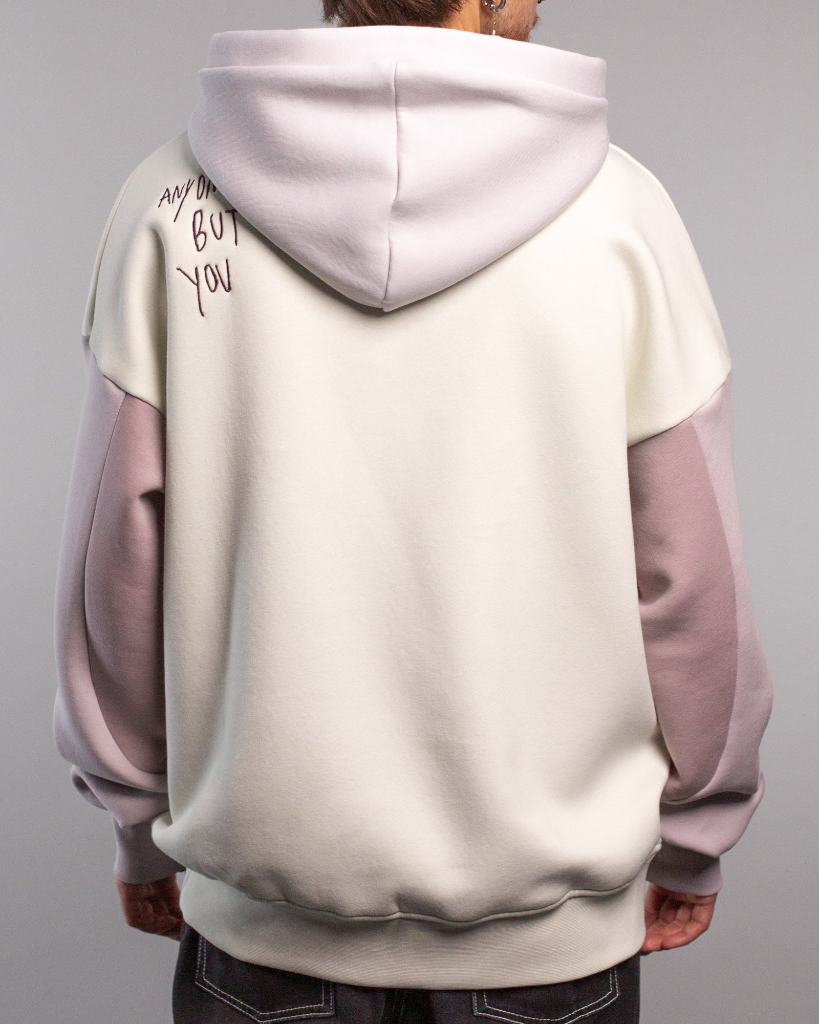 Rückansicht des Hoodies mit Fokus auf die Kapuze und den Schriftzug „ANYONE BUT YOU“ in der linken Schulterpartie. Die Übergänge zwischen den Farben Creme und Flieder sind klar zu erkennen.