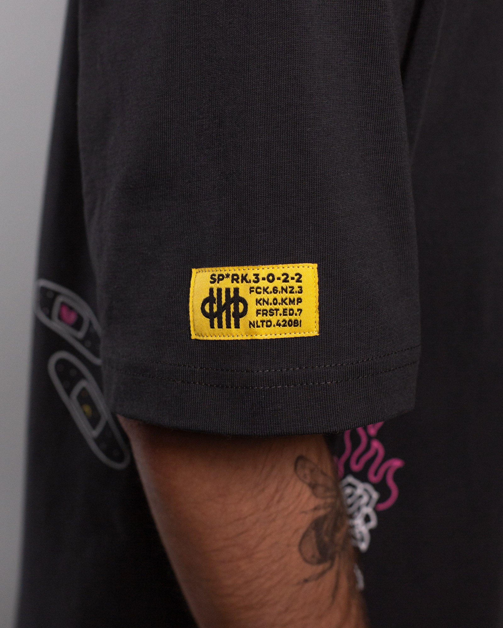 Nahaufnahme des gelben Labelpatches auf dem rechten Ärmel. Die Aufschrift enthält technische Codes und das Logo „SP*RK“. Darunter sind Pflastergrafiken und ein Flammenmotiv zu erkennen.