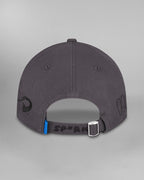 Rückansicht derselben dunkelgrauen Cap. Der hintere Riemen mit silberner Metallschließe trägt die schwarze Aufschrift „SP*RK“. Daneben ist eine kleine blaue Etikettenlasche zu sehen. Auf der linken Seite ist ein geschwungenes schwarzes Symbol aufgestickt, rechts sind stilisierte schwarze Särge mit Gesichtern zu sehen.