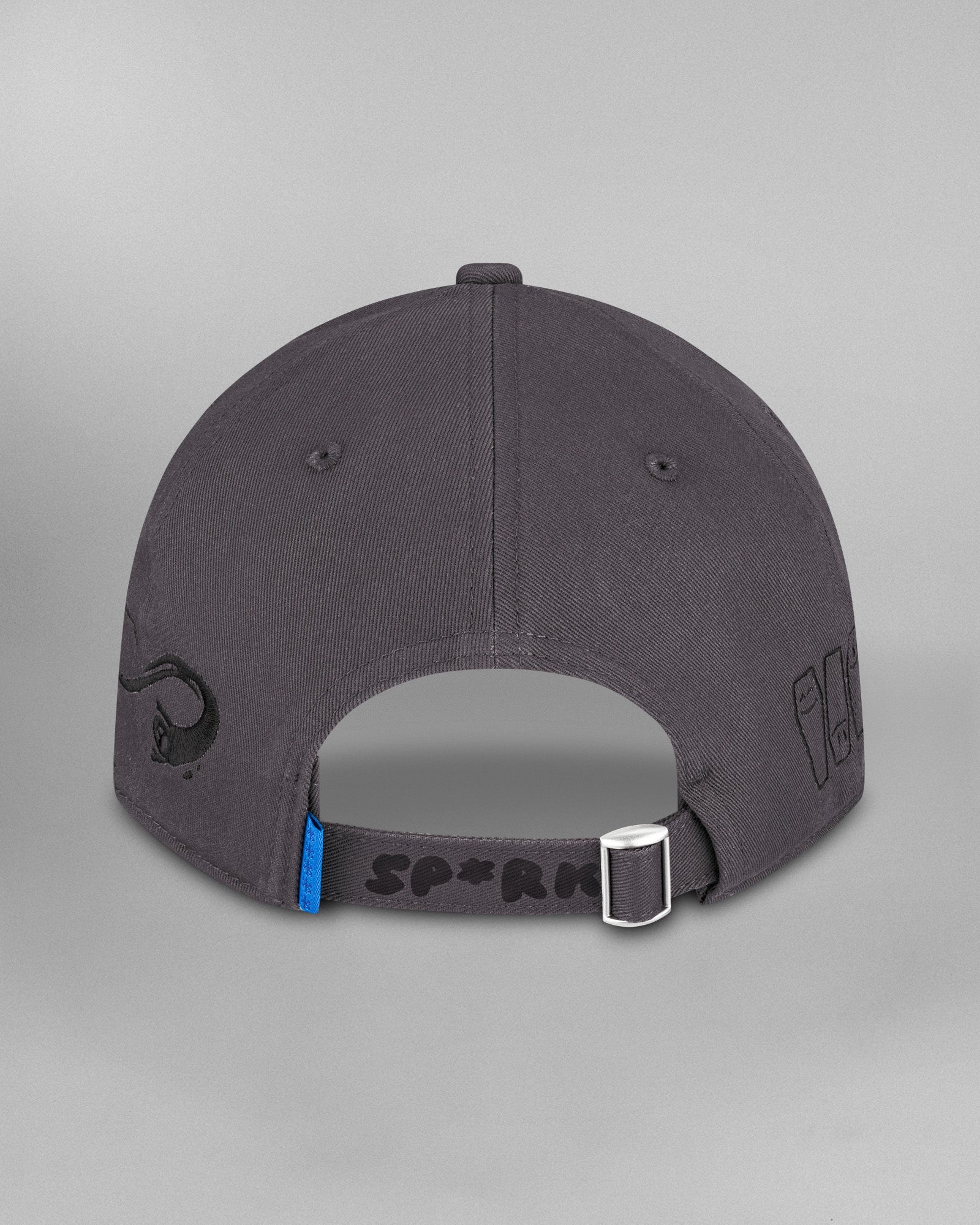 Rückansicht derselben dunkelgrauen Cap. Der hintere Riemen mit silberner Metallschließe trägt die schwarze Aufschrift „SP*RK“. Daneben ist eine kleine blaue Etikettenlasche zu sehen. Auf der linken Seite ist ein geschwungenes schwarzes Symbol aufgestickt, rechts sind stilisierte schwarze Särge mit Gesichtern zu sehen.