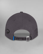Rückansicht derselben dunkelgrauen Cap. Der hintere Riemen mit silberner Metallschließe trägt die schwarze Aufschrift „SP*RK“. Daneben ist eine kleine blaue Etikettenlasche zu sehen. Auf der linken Seite ist ein geschwungenes schwarzes Symbol aufgestickt, rechts sind stilisierte schwarze Särge mit Gesichtern zu sehen.