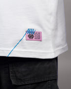 Nahaufnahme des pinkfarbenen Weblabels mit schwarzem Druck und auffälliger blauer Ziernaht am unteren rechten Saum des weißen Shirts. Die Codes auf dem Etikett entsprechen dem Rückenprint.