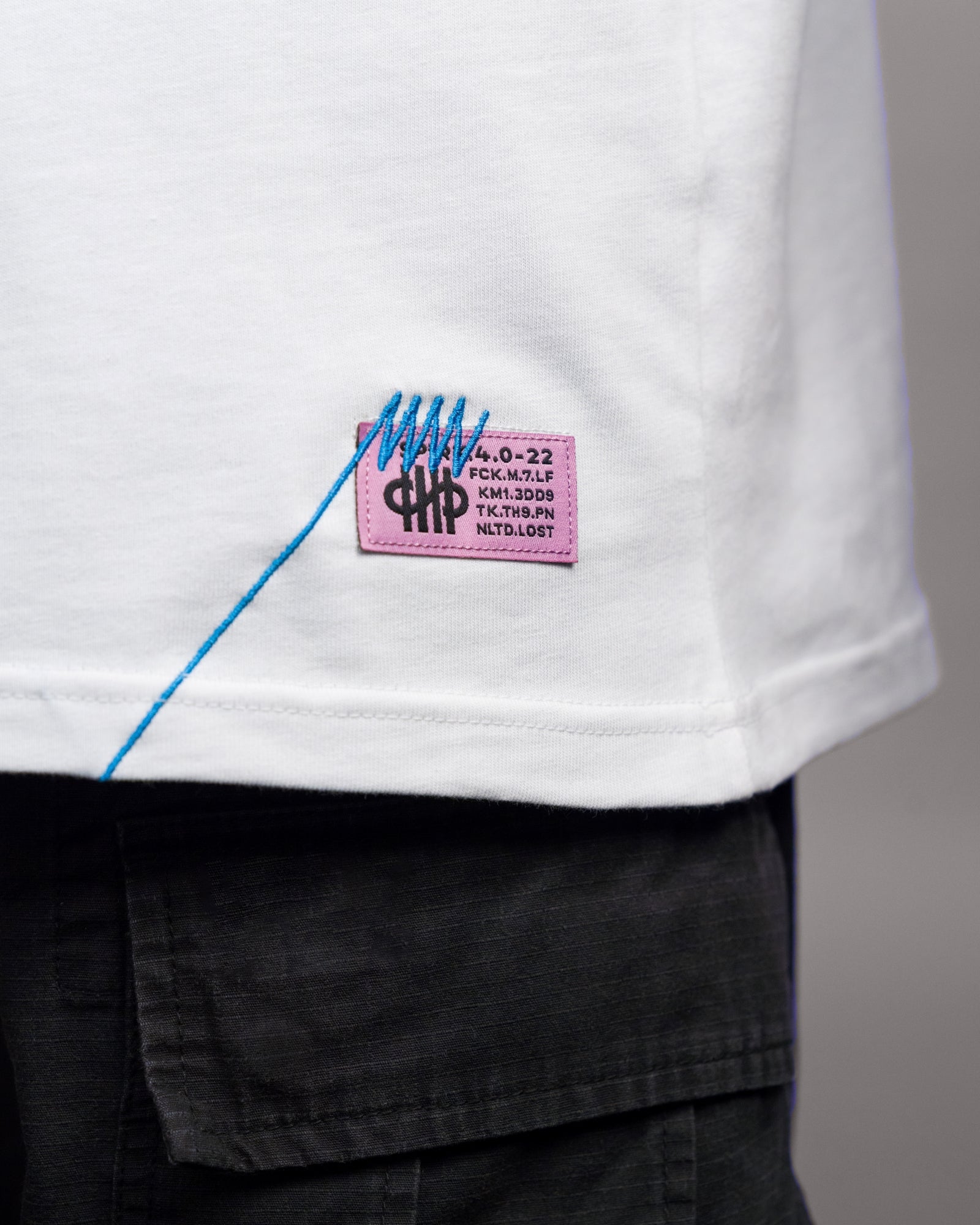 Nahaufnahme des pinkfarbenen Weblabels mit schwarzem Druck und auffälliger blauer Ziernaht am unteren rechten Saum des weißen Shirts. Die Codes auf dem Etikett entsprechen dem Rückenprint.