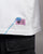 Nahaufnahme des pinkfarbenen Weblabels mit schwarzem Druck und auffälliger blauer Ziernaht am unteren rechten Saum des weißen Shirts. Die Codes auf dem Etikett entsprechen dem Rückenprint.