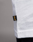 Kleines schwarzes Stoffetikett mit gelbem SP*RK-Schriftzug an der rechten Seitennaht des weißen T-Shirts. Das Etikett ist flach eingenäht.

