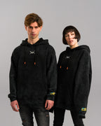 Zwei Personen tragen schwarze Hoodies mit Washed-Effekt. Der Hoodie zeigt auf der Brust ein weißes, stilisiertes Symbol, vorne eine Kängurutasche und an der Ärmelkante ein gelbes Etikett mit blauen Nähten. Die Kapuzenkordeln haben orangefarbene Enden.