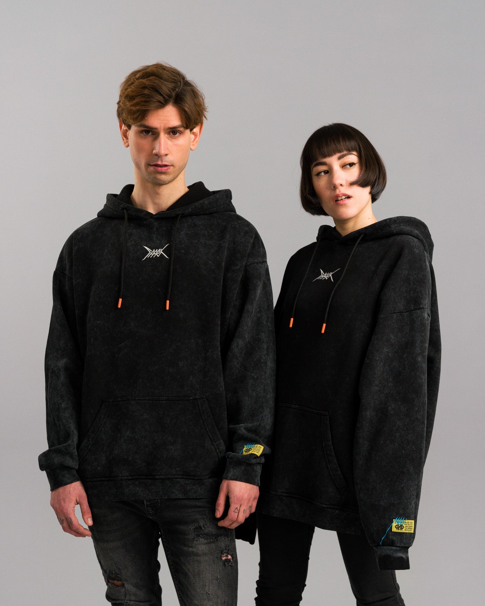 Zwei Personen tragen schwarze Hoodies mit Washed-Effekt. Der Hoodie zeigt auf der Brust ein weißes, stilisiertes Symbol, vorne eine Kängurutasche und an der Ärmelkante ein gelbes Etikett mit blauen Nähten. Die Kapuzenkordeln haben orangefarbene Enden.
