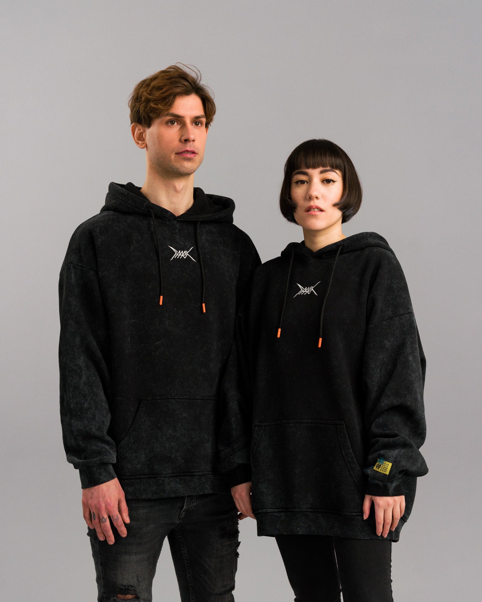 Das Paar blickt nach vorn. Der Hoodie erscheint frontal und locker fallend, mit betonter Bruststickerei, farbigen Details und sichtbarer Labelkante.