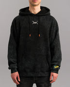 Frontalansicht einer Person mit schwarzem Hoodie. Deutlich sichtbar sind das weiße Symbol auf der Brust, orangefarbene Kordelenden, die Kängurutasche und das gelbe Ärmelpatch mit blauer Naht.