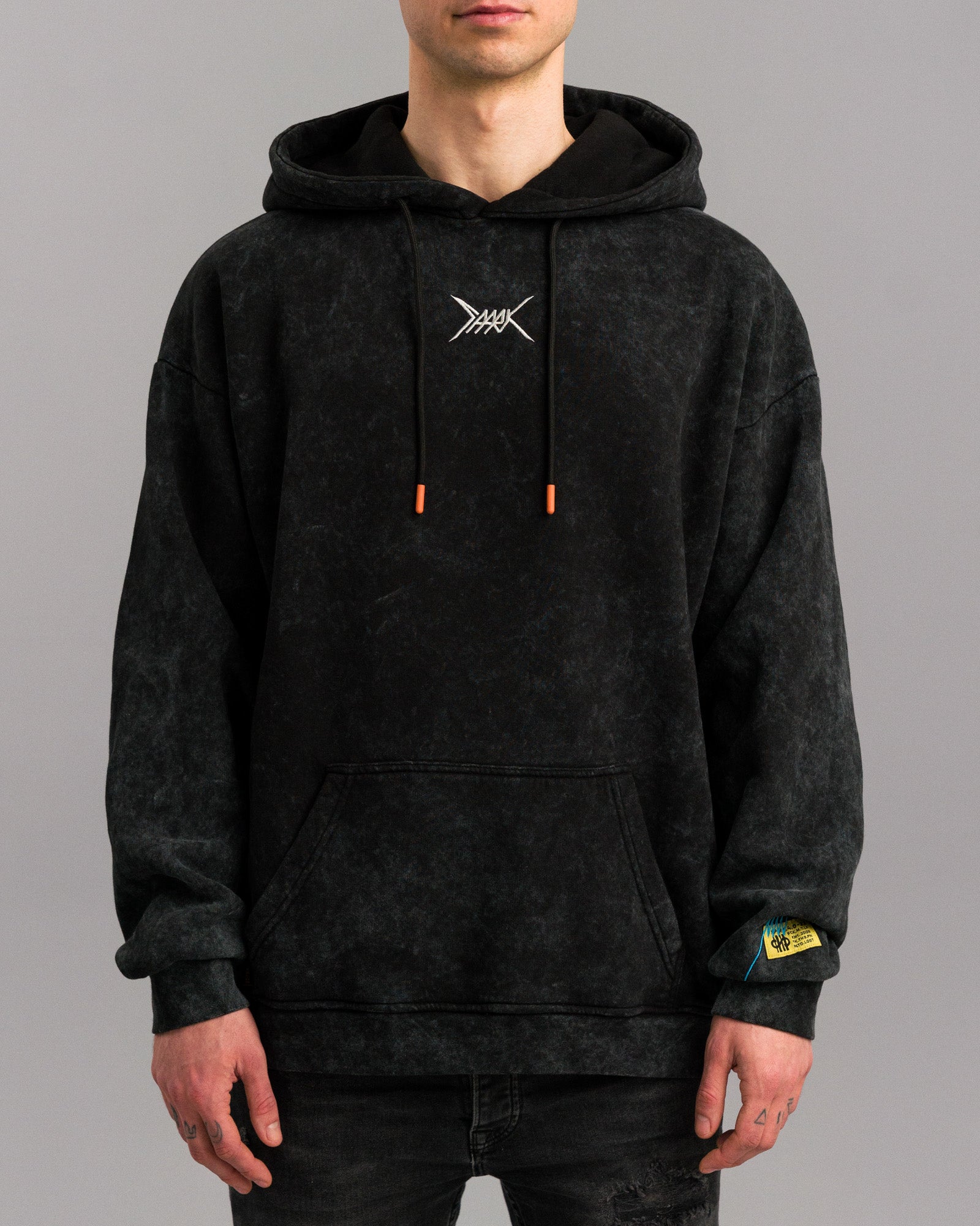 Frontalansicht einer Person mit schwarzem Hoodie. Deutlich sichtbar sind das weiße Symbol auf der Brust, orangefarbene Kordelenden, die Kängurutasche und das gelbe Ärmelpatch mit blauer Naht.