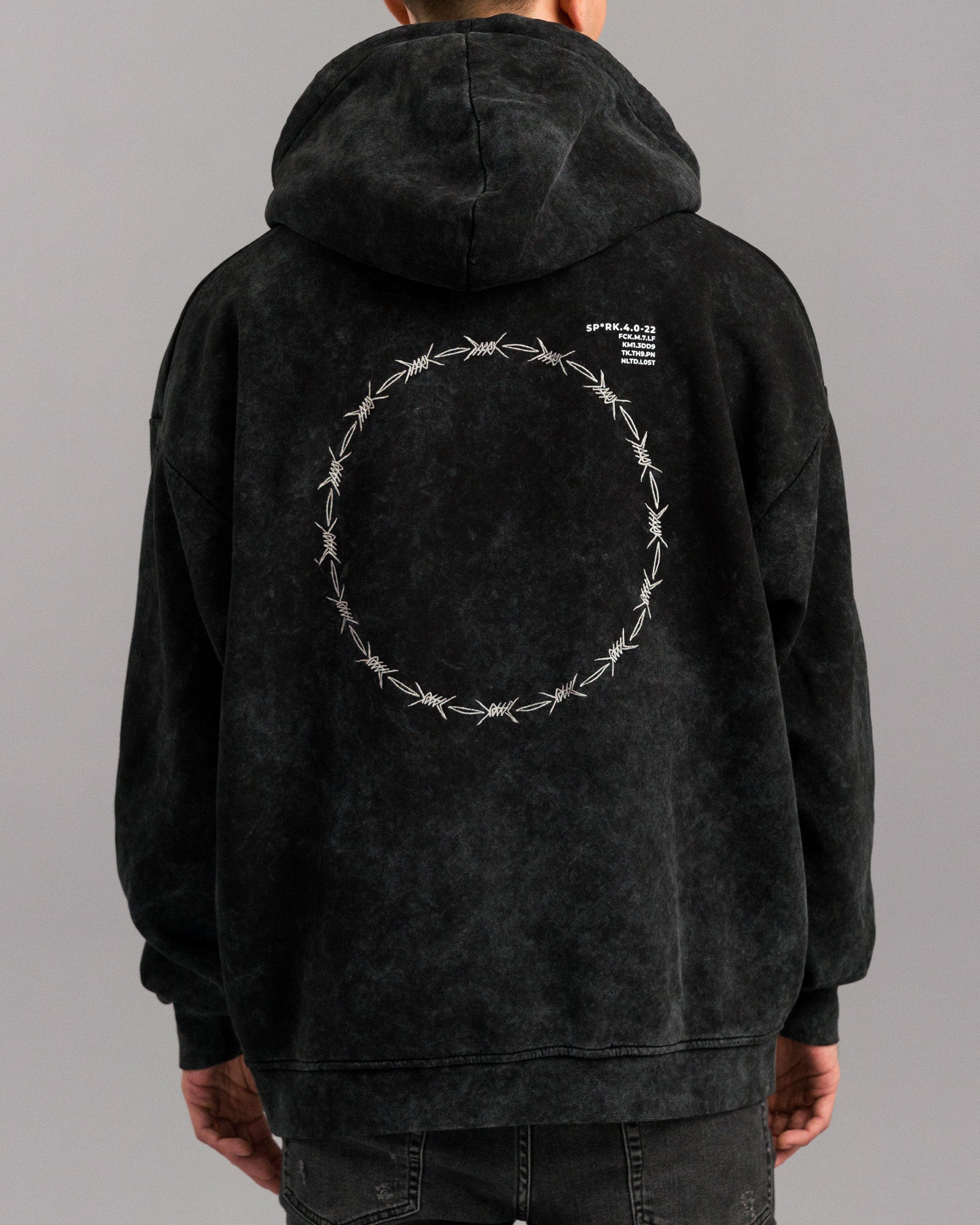  Rückansicht des Hoodies mit großem Rundmotiv aus Stacheldraht-Stickerei auf dem Rücken. Oben rechts auf dem Rücken befinden sich weiße Textdetails mit Code-Zeilen.