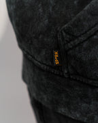 Detailansicht des kleinen schwarzen Labels mit orangefarbener Aufschrift „SP*RK“ an der Seitennaht unter der Kängurutasche des Hoodies.