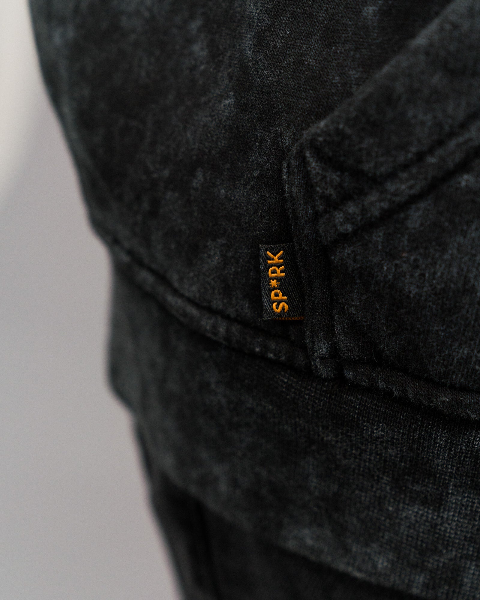 Detailansicht des kleinen schwarzen Labels mit orangefarbener Aufschrift „SP*RK“ an der Seitennaht unter der Kängurutasche des Hoodies.