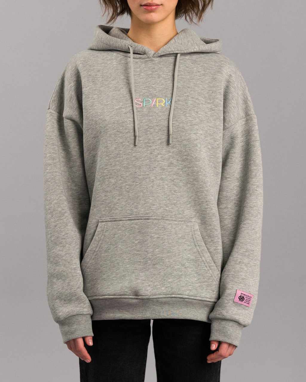 Grauer Kapuzenpullover in lockerer Passform mit Kängurutasche und verstellbarer Kapuze. Auf der Brust ist der Schriftzug „SP/RK“ in bunten Pastellfarben gestickt. Am Ärmelabschluss befindet sich ein kleines rosa Label mit schwarzem Text und grafischen Elementen.