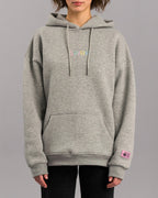 Grauer Kapuzenpullover in lockerer Passform mit Kängurutasche und verstellbarer Kapuze. Auf der Brust ist der Schriftzug „SP/RK“ in bunten Pastellfarben gestickt. Am Ärmelabschluss befindet sich ein kleines rosa Label mit schwarzem Text und grafischen Elementen.