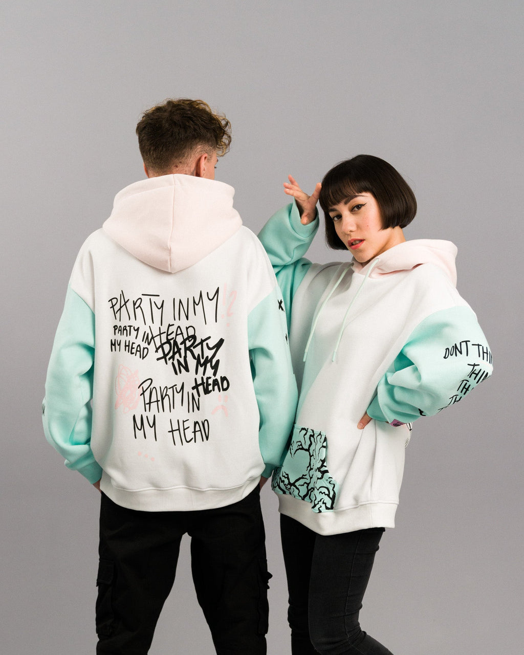 Beide Models in dynamischer Pose. Die Rückseite des linken Hoodies zeigt den „PARTY IN MY HEAD“-Print, der rechte Hoodie ist leicht seitlich gedreht und zeigt Vorderseite mit Tasche und bedrucktem Ärmel.