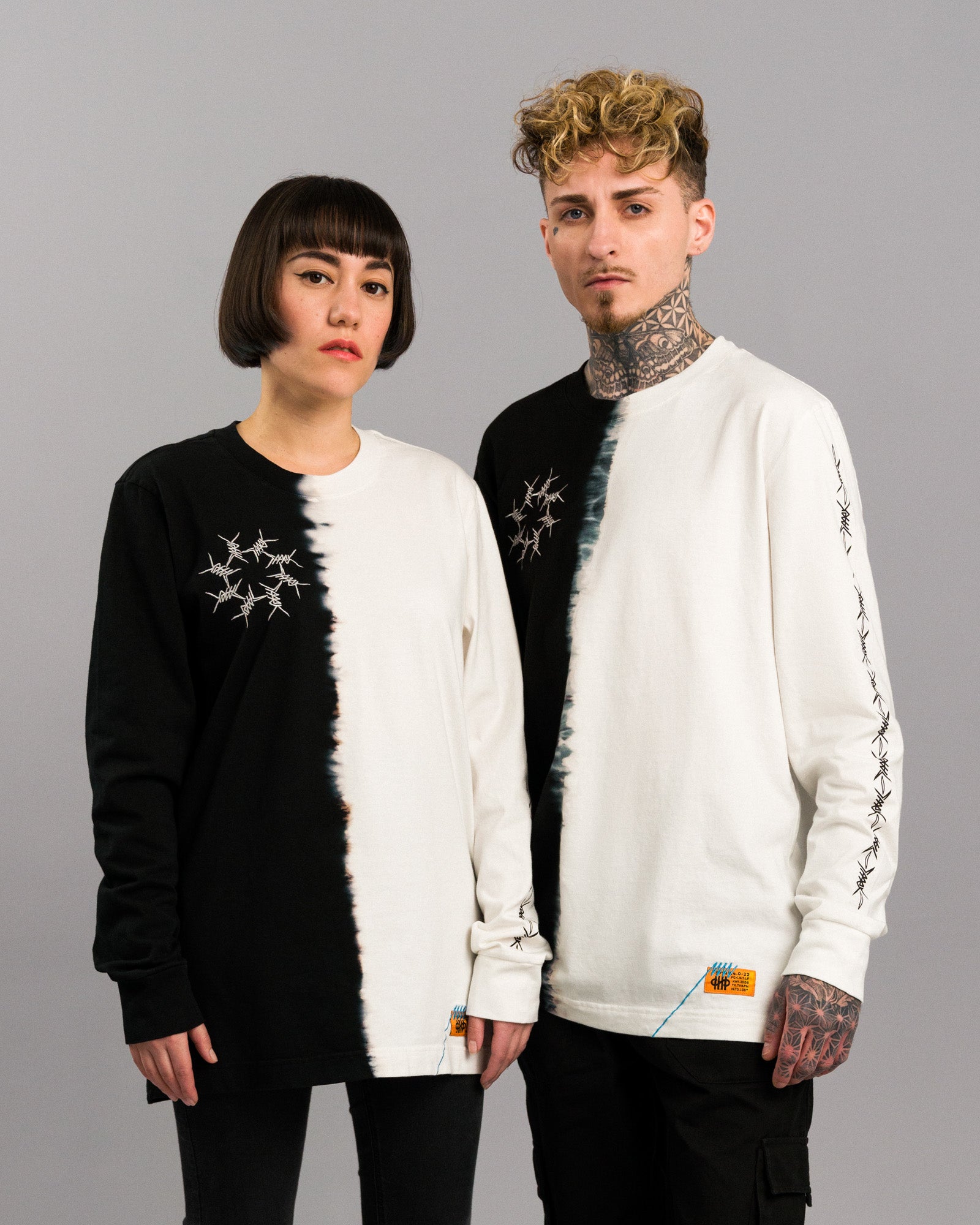 Zweifarbiges Longsleeve mit vertikaler Teilung in Schwarz und Naturweiß. Die Farbkante in der Mitte wirkt unregelmäßig verlaufend im Tie-Dye-Stil. Auf der linken Brustseite ist ein weißes Stacheldraht-Motiv aufgestickt. Das Modell links trägt das Shirt in Schwarz links, das Modell rechts in Weiß links. Beide Shirts zeigen am Saum ein kleines orangefarbenes Patch mit blauer Ziernaht.