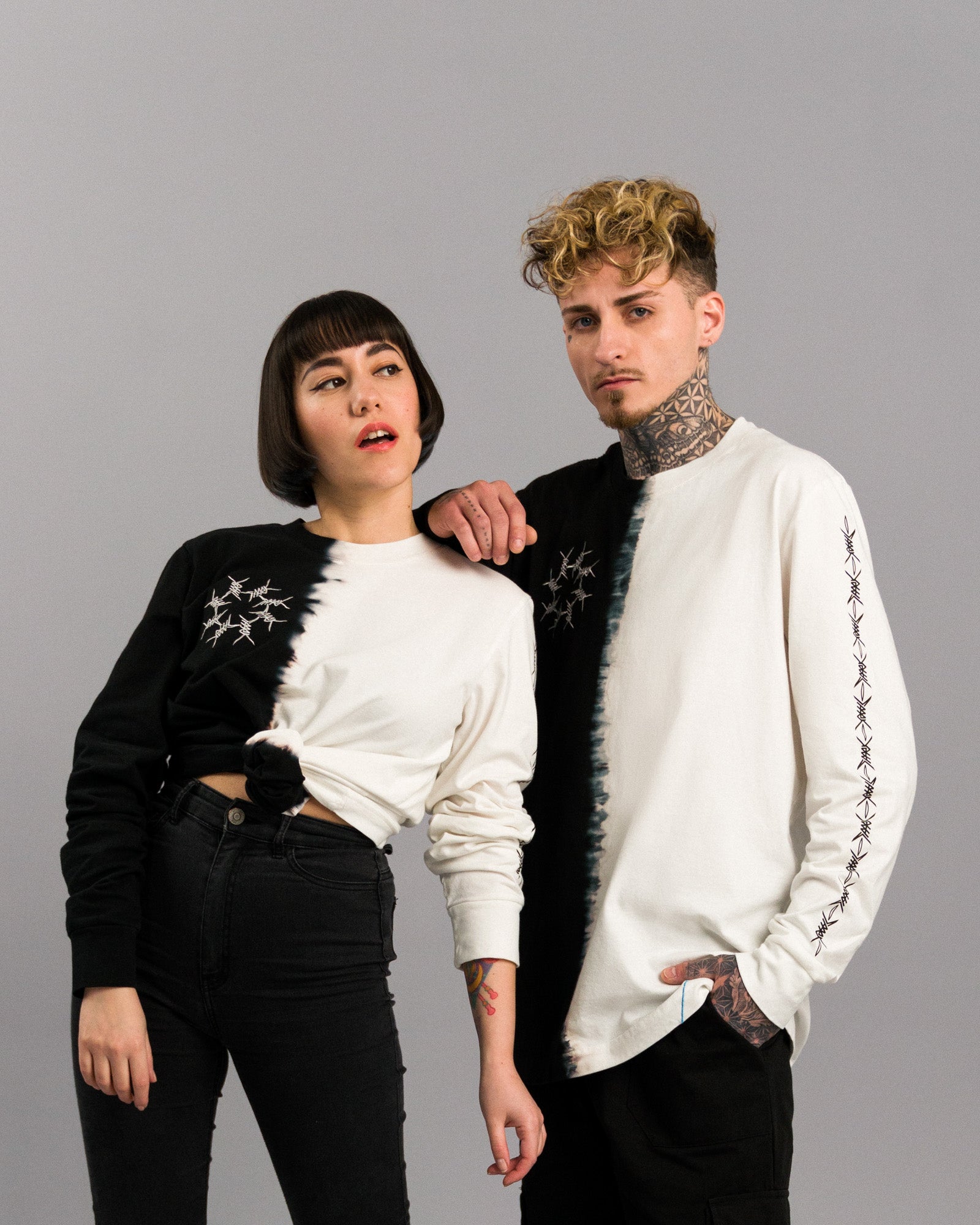 Das Modell links trägt das Shirt mit Knoten in der Taille und hebt die Schulter an. Die linke Shirtseite ist schwarz, die rechte weiß. Das Modell rechts trägt die invertierte Farbvariante. Beide Oberteile zeigen die zentrale Farbtrennlinie, das Stacheldraht-Motiv auf der Brust und das auffällige Label am Saum.